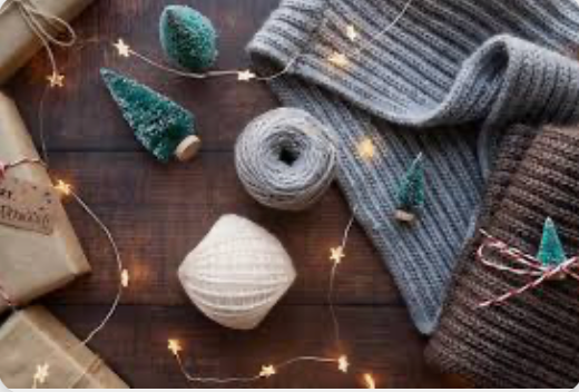 Knitted items and yarn on a wooden surface with string lights and small Christmas trees.
